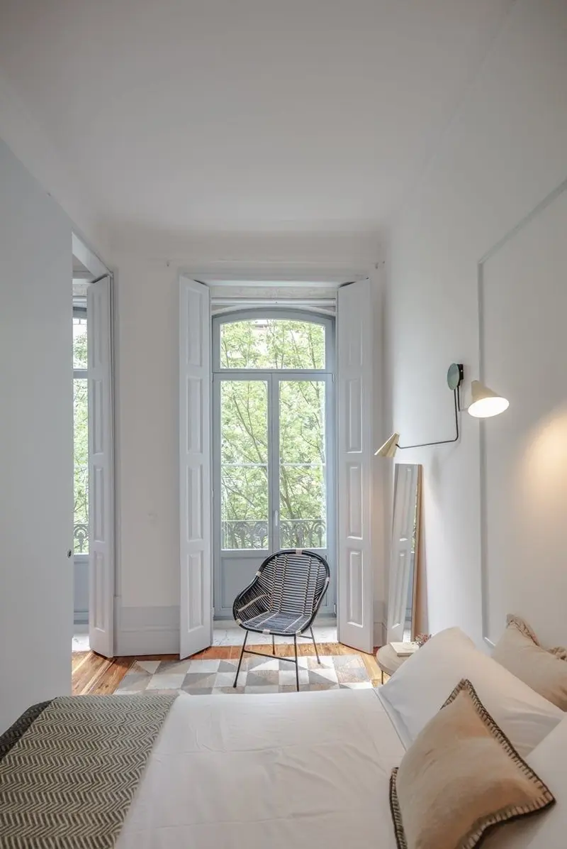 Світла мінімалістична спальня з великими вікнами у підлогу, плетеним кріслом та геометричним килимком